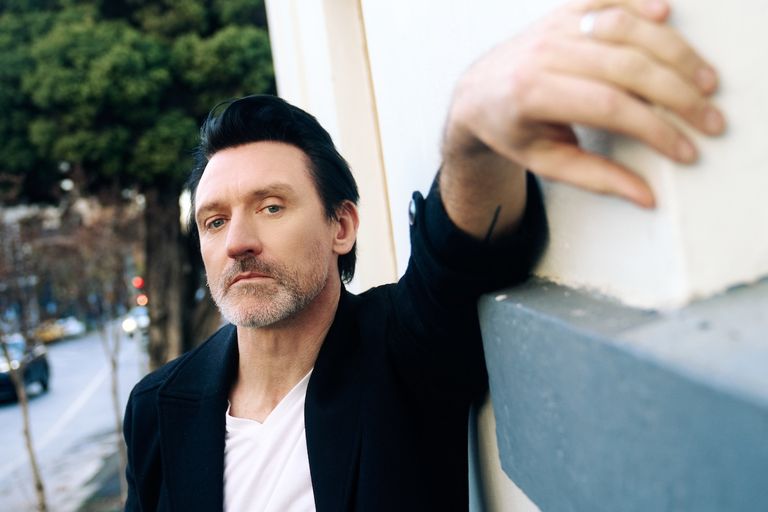 Australian musician Paul Dempsey leaning against a wall outdoors, wearing a black jacket and white t-shirt, looking directly at the camera.